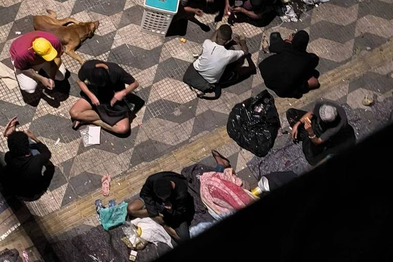 A imagem mostra um grupo de pessoas sentadas em uma calçada, algumas delas estão deitadas e outras estão em atividades diversas. O chão é coberto por um padrão de pedras. Há também um cachorro próximo a um dos grupos. Algumas pessoas estão usando roupas escuras e outras têm roupas coloridas. 