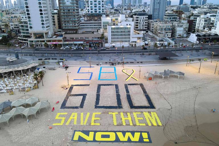 A imagem mostra uma grande mensagem escrita na areia de uma praia, com os números '600' e a frase 'SAVE THEM NOW!' em letras grandes e coloridas. O fundo é composto por prédios altos e uma vista urbana, com um céu nublado. A mensagem é destacada em amarelo e azul, e há um laço amarelo, simbolizando apoio a uma causa.