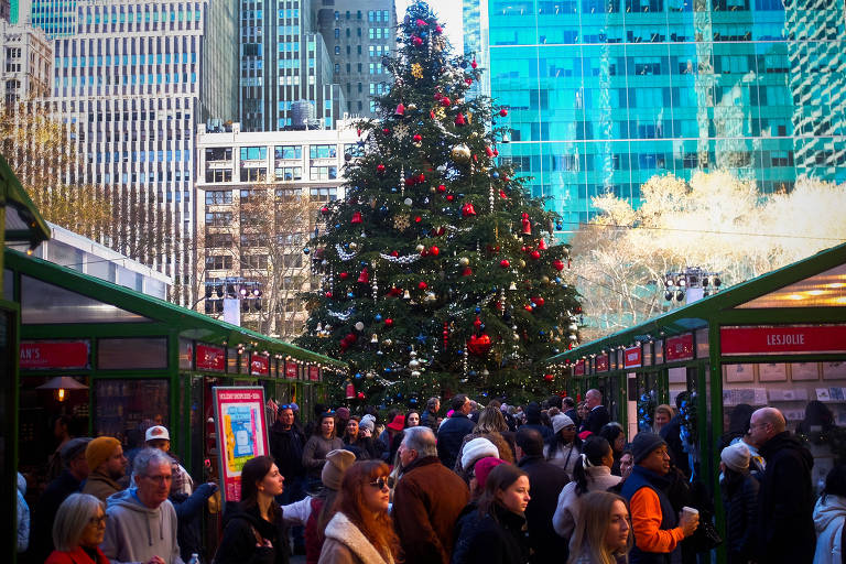 A imagem mostra uma grande árvore de Natal decorada com luzes e enfeites vermelhos e dourados, localizada em um mercado ao ar livre. Há muitas pessoas ao redor, algumas paradas e outras caminhando, em um ambiente urbano com prédios altos ao fundo. O céu está claro e a atmosfera é festiva, típica da época natalina.