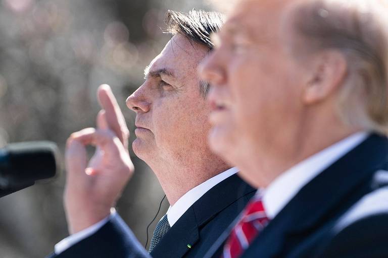 A imagem mostra dois homens em um evento ao ar livre. O homem à esquerda está de perfil, com a mão levantada, enquanto o homem à direita está em foco, mas parcialmente desfocado. Ambos parecem estar em um momento de discurso ou apresentação, com microfones à frente. O fundo é desfocado, sugerindo um ambiente de conferência ou coletiva de imprensa.