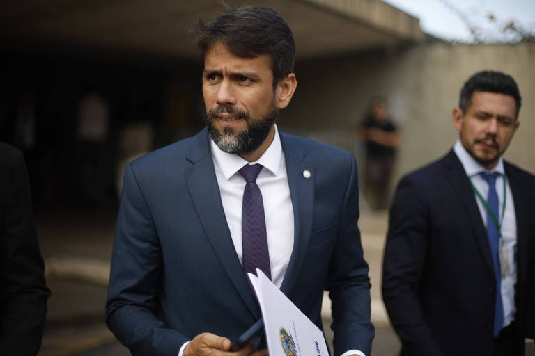 Um homem com cabelo escuro e barba, vestido com um terno azul escuro e uma gravata roxa, está segurando documentos em uma mão. Ele parece estar em um ambiente externo, com uma estrutura de concreto ao fundo. Ao lado dele, há outro homem vestido de forma semelhante, com um crachá visível. O homem à esquerda parece estar em uma conversa ou prestando atenção em algo.