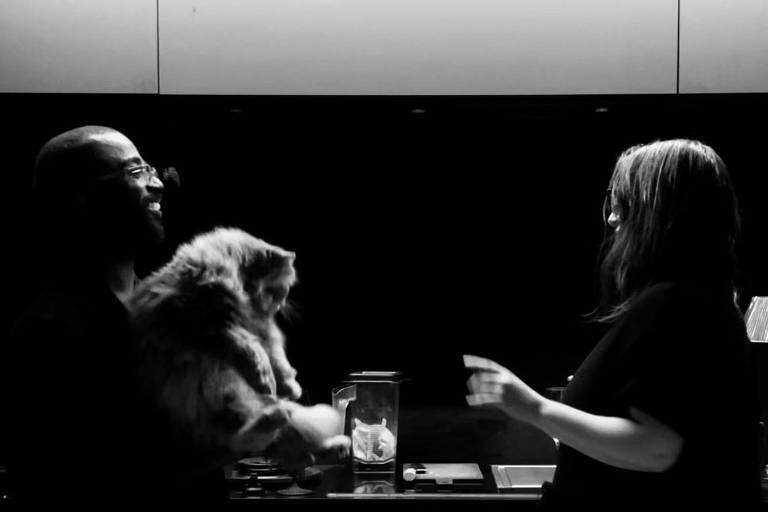A imagem em preto e branco mostra duas pessoas em um ambiente interno. Uma delas está segurando um gato, enquanto a outra está de pé, gesticulando com a mão. O fundo é escuro, com uma superfície que parece ser uma bancada, onde há um copo com gelo. A iluminação é suave, destacando as silhuetas das pessoas e do gato.