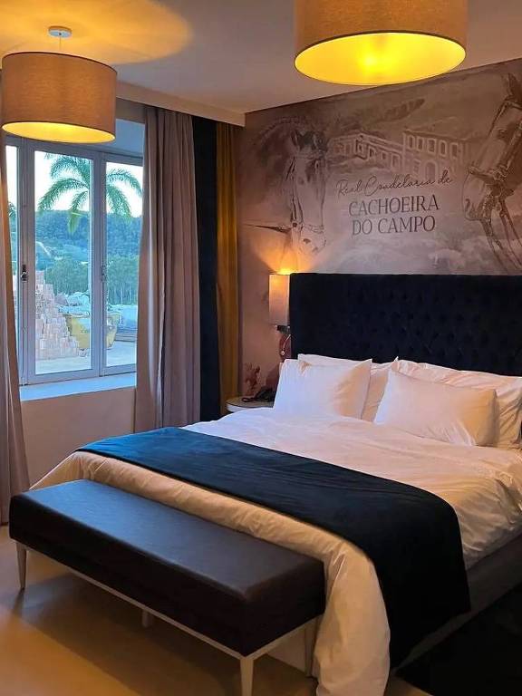 Una moderna habitación de hotel con una cama grande y cómoda cubierta con sábanas blancas y un edredón oscuro. Hay un banco al pie de la cama y las paredes están decoradas con un papel pintado con una ilustración. La habitación está iluminada por dos luces de techo y una ventana que muestra palmeras en el exterior, lo que sugiere un clima tropical.