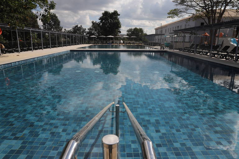 La imagen muestra una piscina con azulejos azules, que reflejan el cielo nublado y los árboles circundantes. En el borde de la piscina hay pasamanos metálicos y al fondo se ven tumbonas y sombrillas. El entorno parece ser el de un complejo turístico o de un área de ocio.