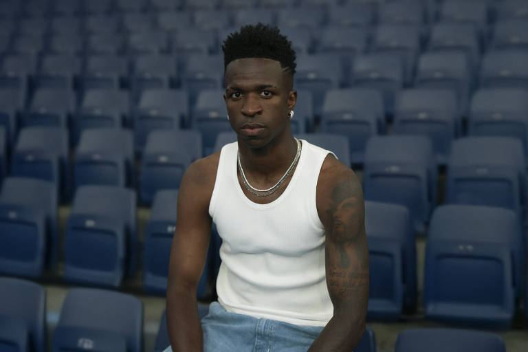 Um homem jovem está sentado em uma fileira de assentos vazios em um estádio. Ele usa uma camiseta sem mangas branca e calças jeans claras. O homem tem cabelo curto e encaracolado, e está olhando diretamente para a câmera com uma expressão séria. O fundo é composto por assentos azuis dispostos em fileiras.