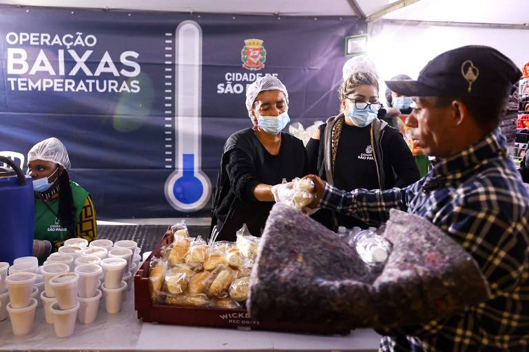 A imagem mostra um evento de apoio a pessoas em situação de vulnerabilidade durante baixas temperaturas. No fundo, há uma faixa com a inscrição 'OPERAÇÃO BAIXAS TEMPERATURAS' e o brasão da cidade de São Paulo. À frente, duas mulheres, usando máscaras e toucas, estão distribuindo alimentos e bebidas quentes para um homem que está recebendo um pacote. Há copos descartáveis e pães em uma mesa.