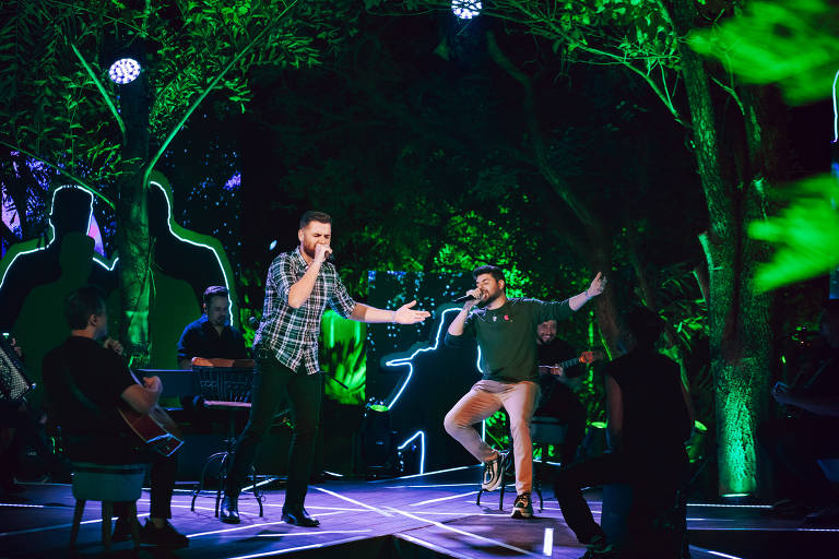 A imagem mostra uma apresentação musical ao ar livre, com dois cantores em destaque. Um deles está vestido com uma camisa xadrez e o outro com um suéter, ambos cantando em um palco iluminado por luzes verdes. Ao fundo, há músicos tocando instrumentos, e a cena é cercada por árvores e uma iluminação que destaca as silhuetas de pessoas. O ambiente é vibrante e festivo, sugerindo um evento musical animado.