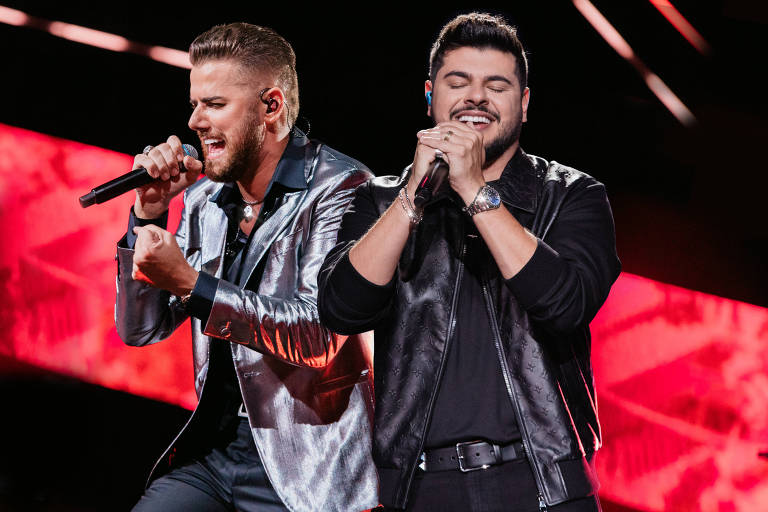 Dois cantores se apresentam em um palco iluminado. O cantor à esquerda está usando uma jaqueta prateada e canta com intensidade, enquanto o cantor à direita, vestido com uma jaqueta preta, sorri e parece estar apreciando o momento. O fundo é composto por luzes vermelhas e padrões vibrantes.