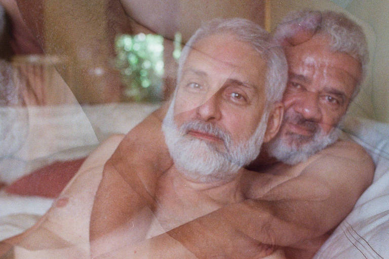 A imagem mostra dois homens com cabelos e barbas grisalhas, nus, em um ambiente de cama. Um homem está sentado na frente, enquanto o outro está atrás, envolvendo-o com os braços. A iluminação é suave, criando uma atmosfera íntima e calorosa. A imagem tem um efeito de sobreposição, dando uma sensação de movimento ou de sonho.