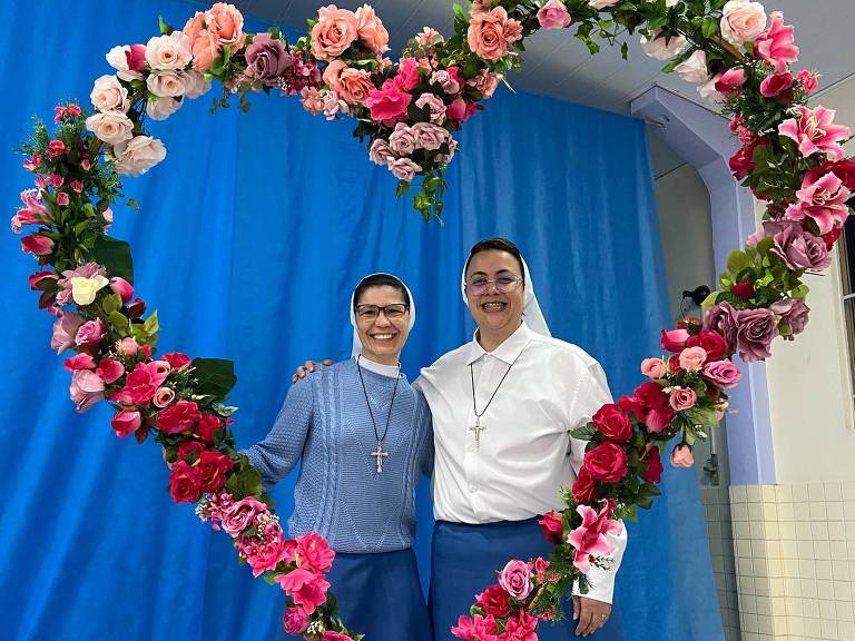 Duas mulheres estão posando para a foto em frente a um fundo azul. Elas estão dentro de um grande coração feito de flores coloridas. A mulher à esquerda usa uma blusa azul e uma saia longa, enquanto a mulher à direita está vestida com uma blusa branca e uma saia longa azul. Acima delas, há um texto que diz 'Um momento especial...' e abaixo, 'Com a ECC'.