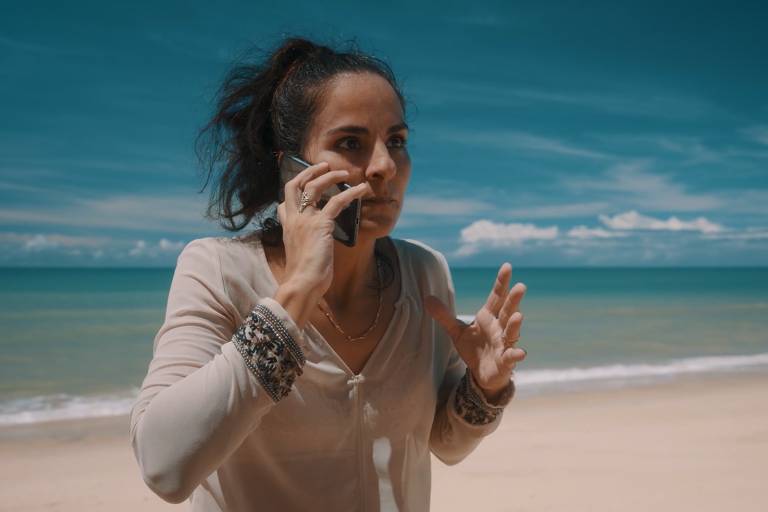Uma mulher está em uma praia, falando ao celular. Ela parece preocupada e gesticula com as mãos. O mar está calmo ao fundo, e o céu é azul com algumas nuvens. A areia da praia é clara e a mulher usa uma blusa de manga longa.