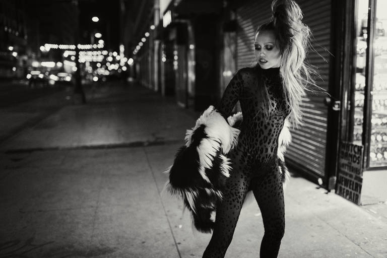 A imagem mostra uma modelo em uma rua à noite, vestindo um macacão de estampa de onça. Ela está posando com uma expressão confiante, segurando uma peça de roupa de pele. O fundo é desfocado, com luzes de rua e lojas visíveis, criando uma atmosfera urbana e noturna. A fotografia é em preto e branco, destacando os contrastes entre a modelo e o ambiente.
