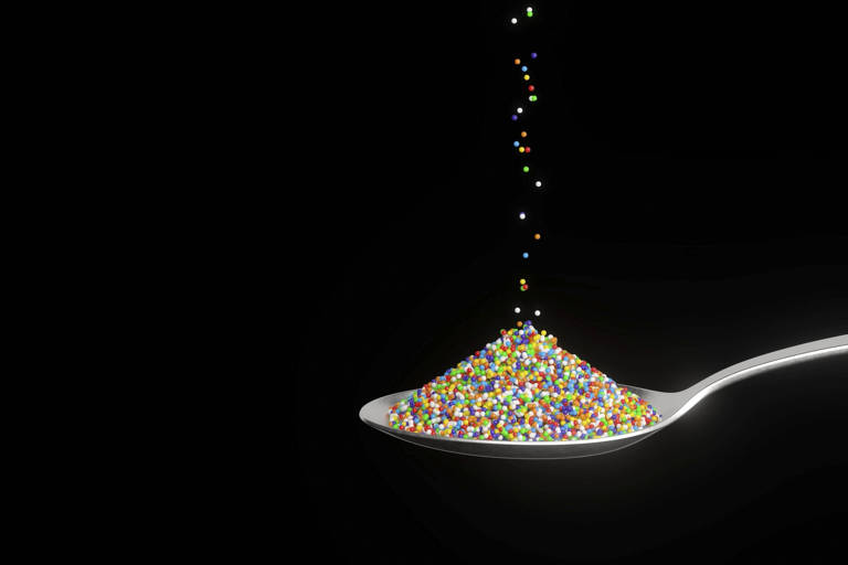 A imagem mostra uma colher prateada cheia de granulado colorido, com pequenos pontos de várias cores. O fundo é preto, destacando a colher e os granulados. Alguns granulados parecem estar caindo da colher, criando um efeito visual dinâmico.