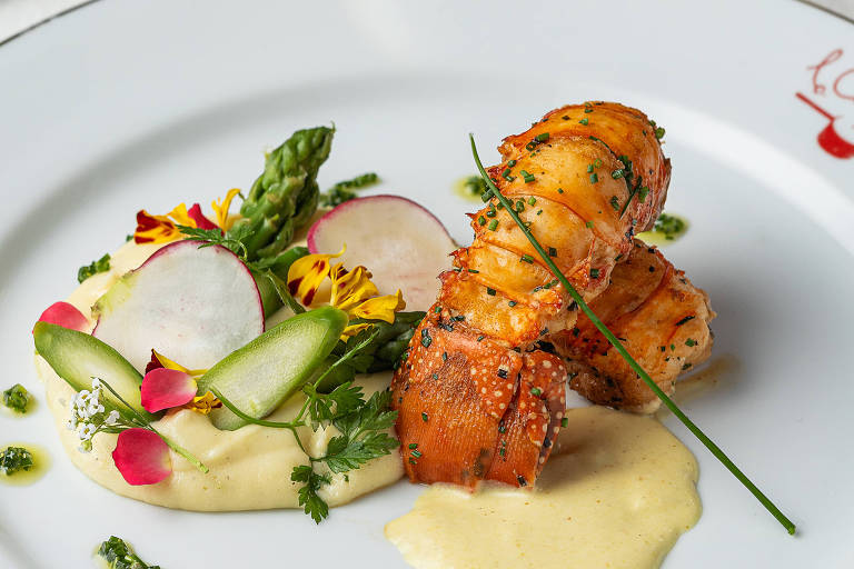 Um prato gourmet apresentado em um prato branco com borda dourada. O prato contém pedaços de lagosta grelhada, acompanhados de purê de batata e uma salada colorida com aspargos, rabanetes e flores comestíveis. Ao fundo, há um talher e uma taça de vinho.