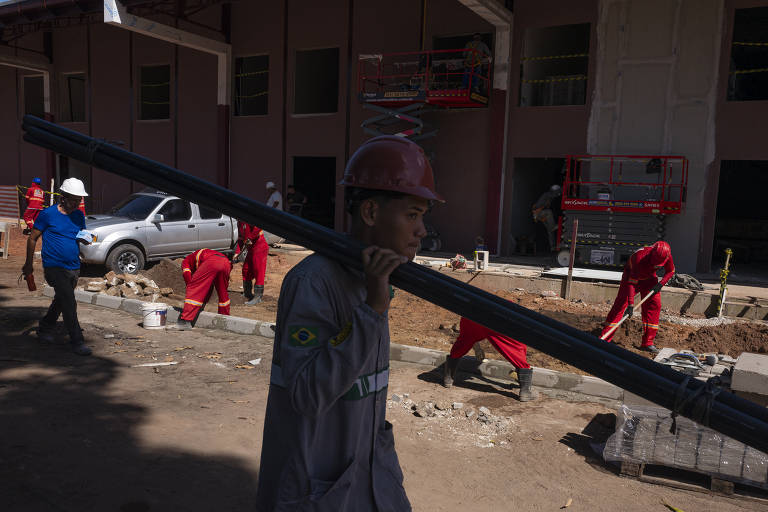 A imagem mostra um trabalhador de construção civil carregando uma barra de metal sobre o ombro. Ele usa um capacete e uma camisa de manga longa. Ao fundo, há outros trabalhadores vestidos de vermelho, alguns usando andaimes e outros cavando no chão. Um veículo está estacionado próximo ao local da obra, e a cena é iluminada pela luz do dia.