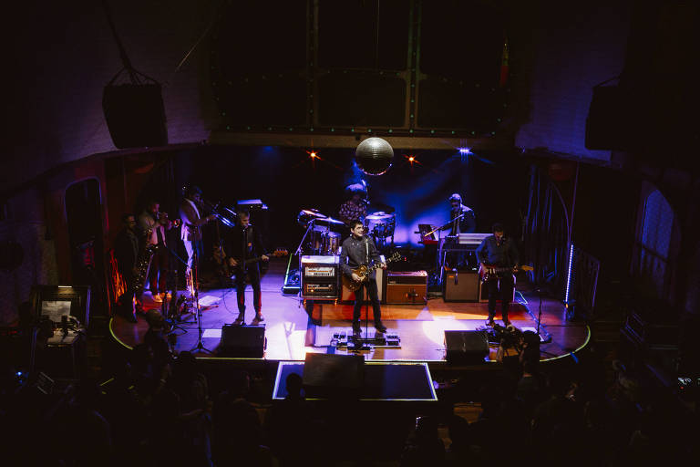 A imagem mostra uma banda se apresentando em um palco iluminado. Há vários músicos tocando instrumentos, incluindo guitarras e teclados. O palco é decorado com luzes coloridas e um grande disco de espelho pendurado no teto. A plateia é visível na parte inferior da imagem, com algumas pessoas assistindo ao show.