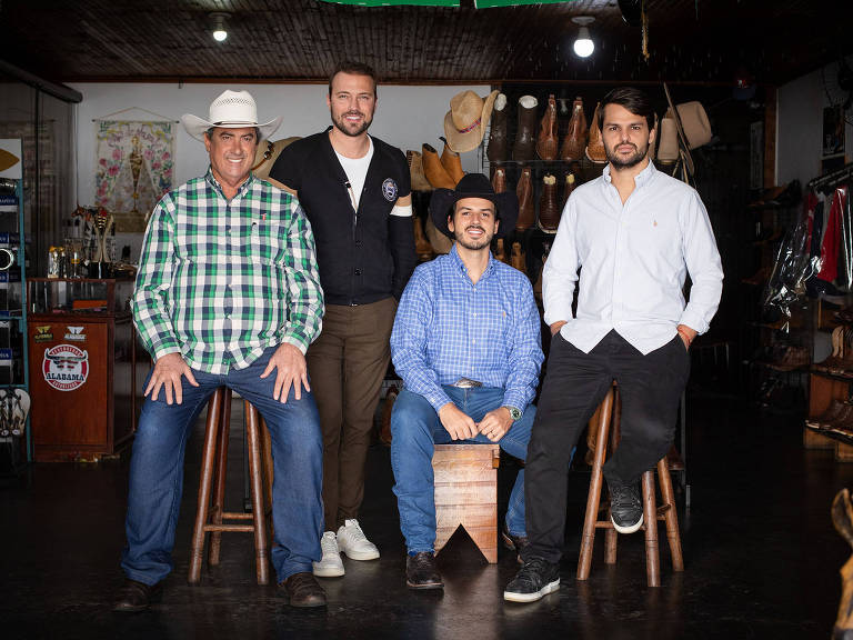 Quatro homens estão posando em uma loja de produtos rurais. O primeiro à esquerda usa uma camisa quadriculada verde e branca, calças jeans e um chapéu branco. O segundo homem, no centro, veste um colete preto sobre uma camisa branca e calças verdes. O terceiro homem usa uma camisa azul xadrez e calças escuras, enquanto o quarto homem à direita está vestido com uma camisa branca e calças escuras. Ao fundo, há prateleiras com produtos e acessórios relacionados ao campo.