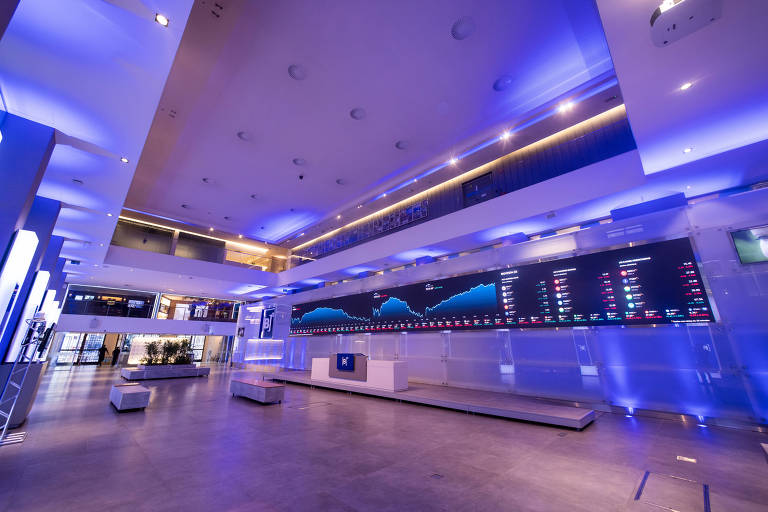 A imagem mostra o interior de um ambiente financeiro moderno, com iluminação azul. No fundo, há uma grande tela exibindo gráficos e dados financeiros. O espaço é amplo, com paredes e piso em tons neutros, e há algumas poltronas brancas dispostas no ambiente.