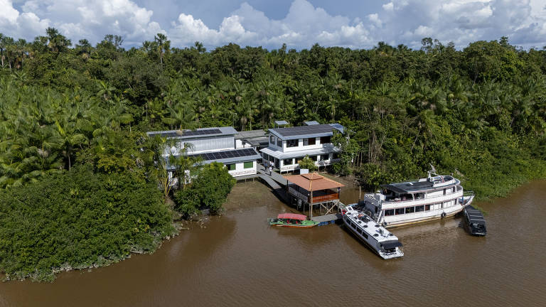 A imagem mostra uma residência moderna localizada à beira de um rio, cercada por vegetação densa. A casa possui um design contemporâneo com várias janelas e varandas. Na frente da casa, há um barco ancorado e uma pequena plataforma de madeira. O rio apresenta águas marrons e a vegetação ao redor é predominantemente verde, com palmeiras e outras plantas tropicais. O céu está parcialmente nublado.