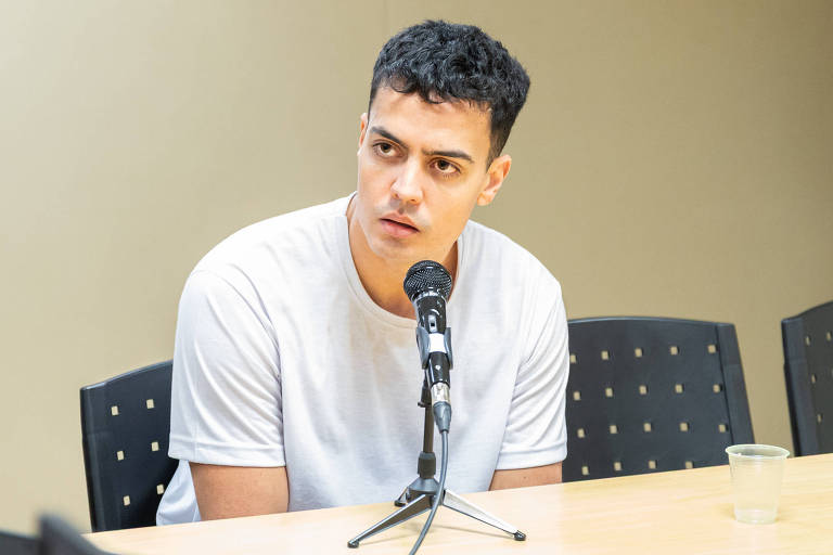 Un jeune homme aux cheveux courts et bouclés, vêtu d'un t-shirt blanc, est assis à une table. Il semble être dans une interview ou une conférence de presse, fixant intensément le microphone devant lui. En arrière-plan, il y a des chaises noires et un verre transparent sur la table.