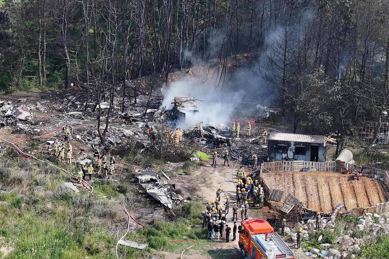 A imagem mostra o local de um acidente aéreo, onde destroços de uma aeronave estão espalhados em uma área que parece ter sido afetada por um incêndio. Há fumaça saindo dos destroços e várias pessoas, possivelmente bombeiros e equipes de resgate, estão presentes na cena. Ao fundo, árvores queimadas e um terreno acidentado são visíveis. Um caminhão de bombeiros está estacionado em primeiro plano, e há uma estrutura de madeira que parece ser uma construção próxima ao local do acidente.