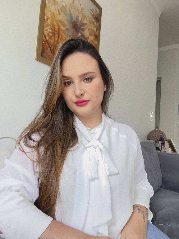 A imagem mostra uma mulher sentada em um sofá cinza, com uma blusa branca e um laço na gola. Ela tem cabelos longos e soltos, e está olhando para a câmera com um leve sorriso. Ao fundo, há uma parede clara e uma pintura decorativa com flores. O ambiente parece bem iluminado e acolhedor.