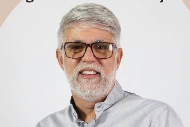 Um homem de cabelos grisalhos e barba, usando óculos e uma camisa de botão clara. Ele está sorrindo e posando para a foto, com um fundo neutro. A imagem tem um design circular ao redor do homem.