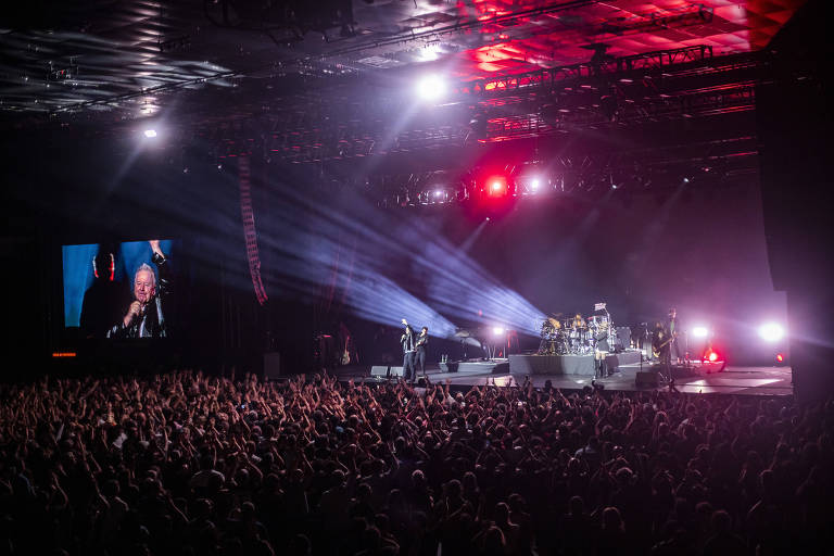A imagem mostra um grande show ao vivo com uma multidão de pessoas levantando as mãos. No palco, há uma banda tocando, com instrumentos visíveis. À esquerda, uma tela grande exibe uma imagem de um dos membros da banda. O ambiente é iluminado por luzes coloridas e há um efeito de fumaça no ar.