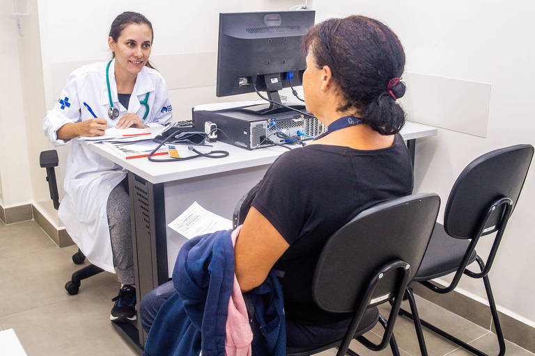 A imagem mostra uma consulta médica em um consultório. À esquerda, uma médica vestindo um jaleco branco e com um estetoscópio ao redor do pescoço está sentada em uma cadeira, olhando para uma paciente. A paciente, que está sentada em uma cadeira oposta, parece estar ouvindo atentamente. Na mesa, há um computador, papéis e um esfigmomanômetro. O ambiente é claro e moderno, com piso de cerâmica e paredes brancas.