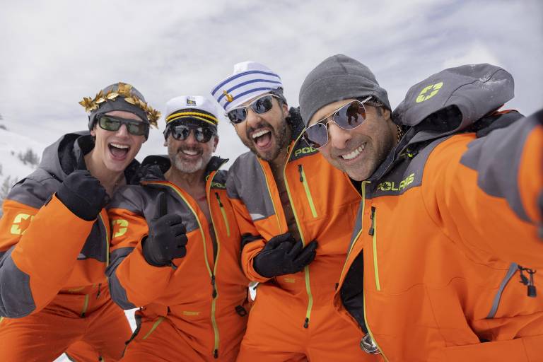 Quatro homens posando para uma selfie em uma área de neve. Eles estão usando roupas de esqui laranja e acessórios divertidos, como óculos escuros e chapéus. O fundo mostra um céu nublado e montanhas cobertas de neve.