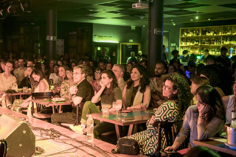 A imagem mostra um público assistindo a uma apresentação em um local de eventos. As pessoas estão sentadas em mesas, algumas com copos e bebidas. O ambiente é iluminado com luzes suaves e há uma grande quantidade de pessoas visíveis, refletindo um clima de entretenimento e socialização. Ao fundo, há uma prateleira com bebidas e uma iluminação que destaca o bar.