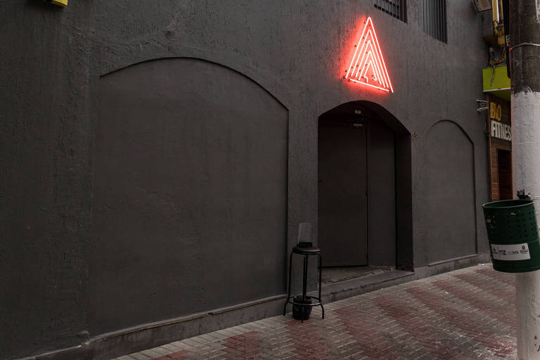 A imagem mostra a fachada de um edifício com paredes cinzas. Há uma iluminação em forma de triângulo vermelho na parte superior central da entrada. As janelas são gradeadas e há um pequeno espaço em frente à entrada, onde está um suporte preto. À esquerda, há um dispositivo amarelo na parede. O calçamento é de pedras em tons de vermelho e cinza, e há uma árvore à esquerda da imagem.