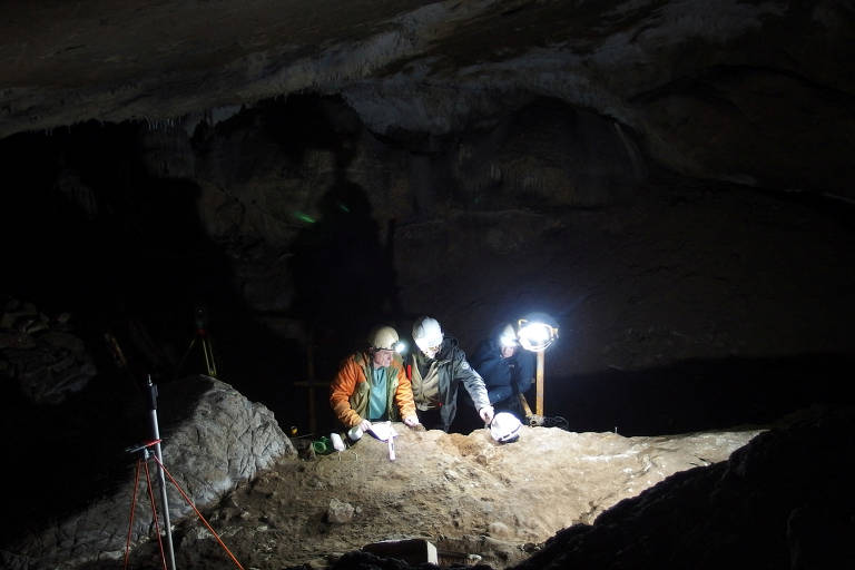 A imagem mostra duas pessoas trabalhando em uma caverna escura, iluminadas por luzes artificiais. Elas estão agachadas sobre o solo, que parece ser de terra e areia, e estão utilizando ferramentas para escavação. Ao fundo, a caverna apresenta formações rochosas e uma iluminação fraca, criando um ambiente de exploração. Há também um tripé com equipamento de medição ou iluminação próximo a elas.