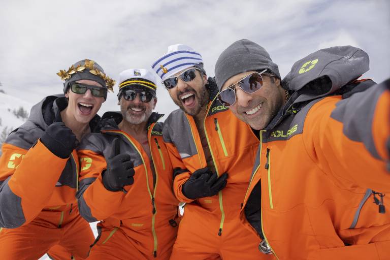 Quatro homens posando para uma selfie em uma área de neve. Eles estão usando roupas de esqui laranja e acessórios divertidos, como óculos escuros e chapéus. O fundo mostra um céu nublado e montanhas cobertas de neve.