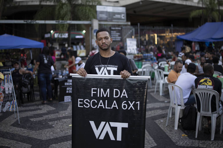 Um homem está em pé segurando um cartaz que diz 'FIM DA ESCALA 6X1' e 'VAT'. Ao fundo, há uma multidão de pessoas sentadas em cadeiras plásticas em um espaço público, com barracas azuis montadas. O ambiente parece ser um local de protesto ou manifestação.