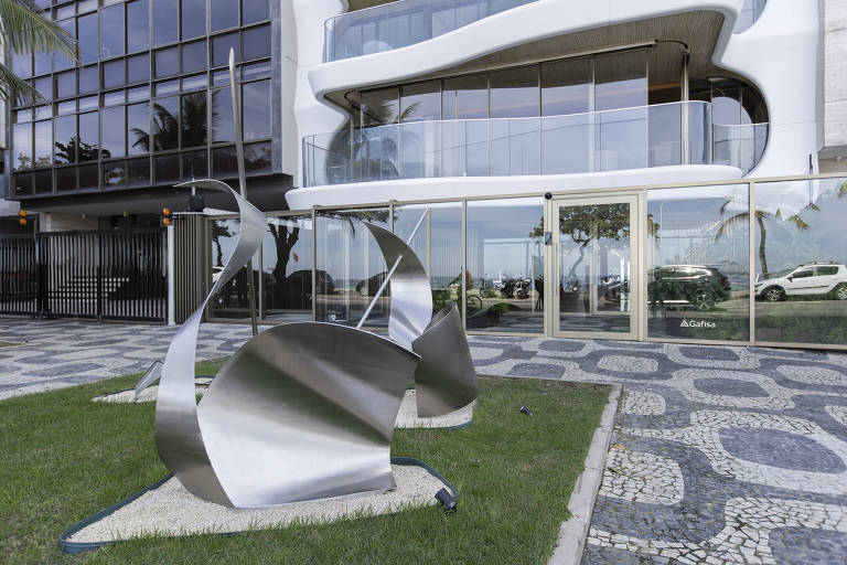 A imagem mostra uma escultura moderna em metal, com formas curvas e polidas, localizada em um gramado em frente a um edifício de vidro. O edifício possui uma fachada contemporânea, com grandes janelas e um design arrojado. Ao fundo, é possível ver árvores e o mar, sugerindo que a cena está em uma área costeira. O piso é composto por pedras em um padrão decorativo.
