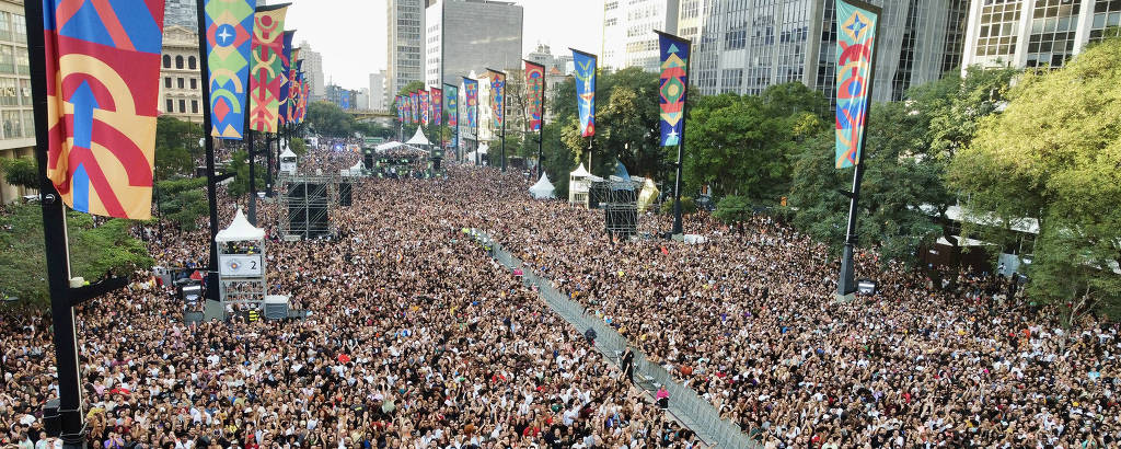 A imagem mostra uma grande multidão reunida em um evento ao ar livre, com pessoas de diversas idades e estilos. O local é cercado por prédios altos e há bandeiras coloridas penduradas ao longo da área. No fundo, pode-se ver um palco e uma estrutura de som, indicando que há uma apresentação acontecendo. A atmosfera parece festiva e animada, com muitas pessoas concentradas em um espaço amplo.