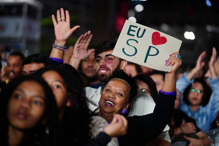Uma multidão em um evento, com pessoas levantando as mãos e sorrindo. Uma mulher na frente segura um cartaz que diz 'EU ❤️ SP'. O ambiente parece festivo, com várias pessoas ao fundo também demonstrando alegria.