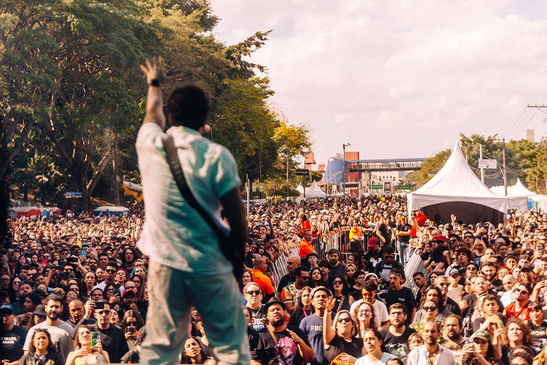 A imagem mostra um grande público em um show ao ar livre. No primeiro plano, um músico de costas, segurando um instrumento, está se apresentando. O público é numeroso, com pessoas de diversas idades e estilos, algumas segurando bandeiras e outras com expressões de entusiasmo. Ao fundo, há árvores e tendas brancas, além de um céu parcialmente nublado.