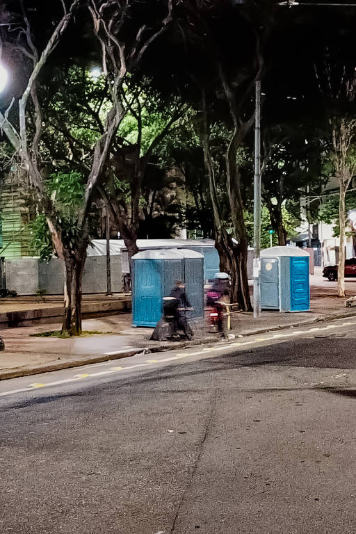 A imagem mostra um cenário noturno em uma rua, onde há banheiros químicos azuis dispostos em uma área com árvores. Duas pessoas estão se deslocando de bicicleta na calçada, enquanto uma lâmpada de rua ilumina a cena. O ambiente parece urbano, com um asfalto visível e uma sinalização de trânsito ao fundo.