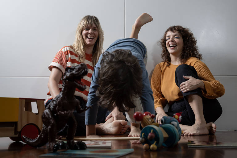 A imagem mostra duas mulheres sorrindo enquanto observam uma criança fazendo uma acrobacia de cabeça para baixo. A criança está no centro da imagem, com o corpo voltado para baixo e as pernas para cima. Ao redor, há brinquedos espalhados pelo chão, incluindo um dinossauro de plástico e outros brinquedos coloridos. O ambiente é interno, com uma parede clara ao fundo e um piso de madeira.