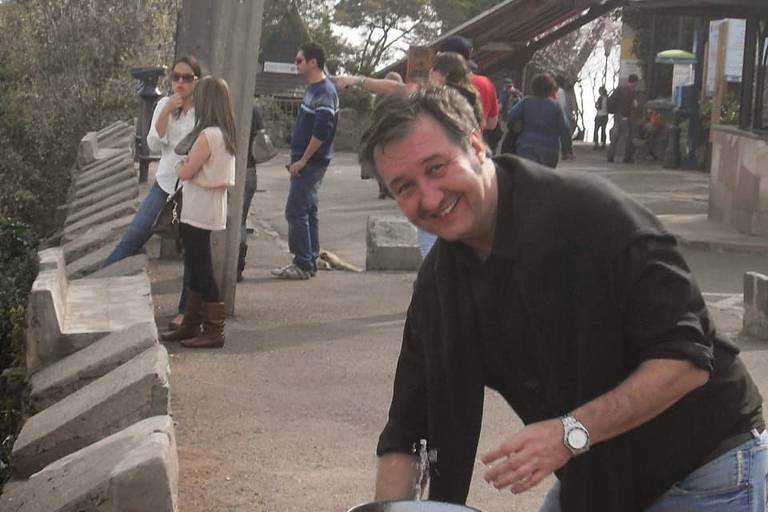 Um homem está sorrindo enquanto se inclina sobre uma fonte de água em um espaço público. Ao fundo, há várias pessoas conversando e se divertindo. O ambiente parece ser um parque ou uma área de lazer, com árvores e um edifício visível. O homem está vestindo uma camisa preta e jeans claros, e a fonte de água está em primeiro plano.