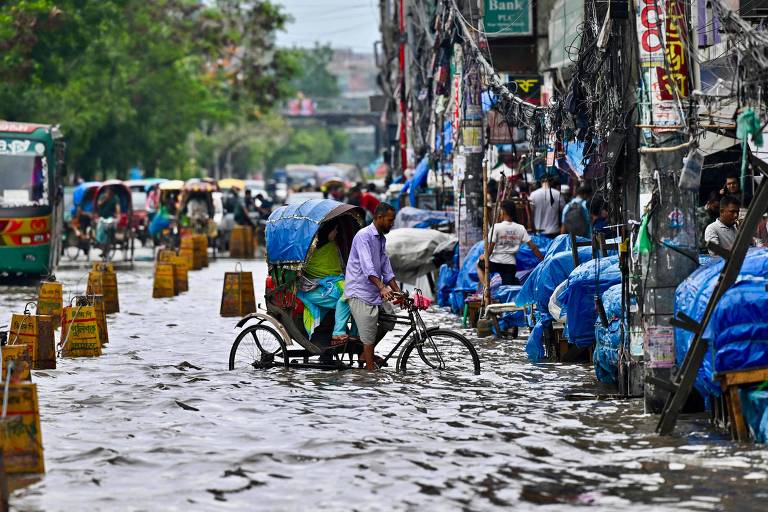 A imagem mostra uma rua inundada em uma área urbana, com água cobrindo a maior parte do chão. Algumas pessoas estão se deslocando, incluindo um homem em uma bicicleta. Ao fundo, há veículos e barracas cobertas com lonas azuis. A vegetação é visível nas laterais, e há fios expostos pendurados entre os prédios.