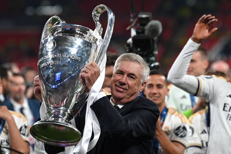 Um homem sorridente segura um troféu grande da Liga dos Campeões da UEFA, enquanto celebra com outros jogadores ao fundo. O homem está vestido com um terno escuro e uma camisa branca, e o troféu brilha sob as luzes do estádio. Outros membros da equipe estão ao redor, alguns levantando os braços em celebração.