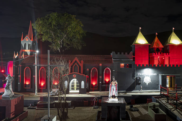 A imagem mostra um castelo iluminado à noite, com torres e telhados em formato pontudo. A estrutura é predominantemente preta com detalhes em vermelho e amarelo. Há uma árvore à esquerda e estátuas em primeiro plano, além de um ambiente que parece ser um parque ou área de lazer. O céu está escuro, sugerindo que a foto foi tirada à noite.