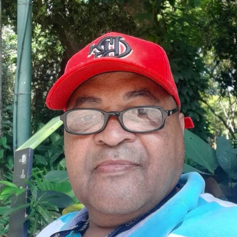 Um homem está posando para a foto em um ambiente natural, cercado por plantas verdes. Ele usa um boné vermelho com um logotipo preto e uma camisa azul. O fundo é composto por árvores e folhagens, sugerindo um dia ensolarado.