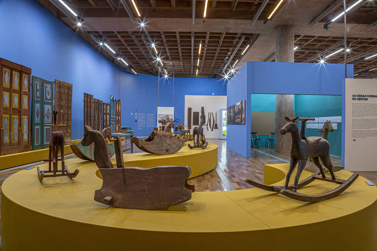 A imagem mostra uma exposição em um museu com várias peças de brinquedos de madeira, incluindo cavalinhos de balanço. As paredes são pintadas de azul e há painéis informativos e obras de arte ao fundo. O espaço é bem iluminado e tem um piso de madeira.