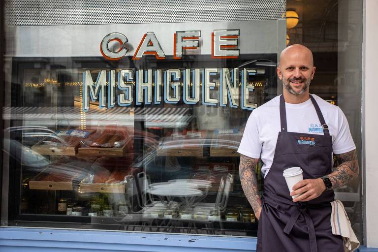 Um homem com tatuagens está em pé em frente a uma vitrine de um café chamado 'Café Mishiguene'. Ele usa uma camiseta branca e um avental escuro, segurando um copo de café. Ao fundo, a vitrine exibe o nome do café em letras grandes e iluminadas, refletindo a movimentação da rua.