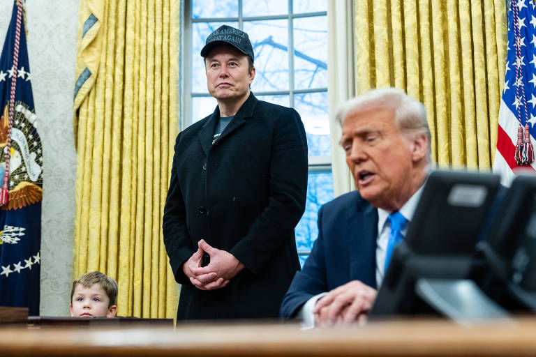 A imagem mostra uma cena no Salão Oval da Casa Branca. À esquerda, um menino pequeno está sentado em uma mesa. Ao centro, Elon Musk está em pé, vestindo um casaco escuro e um boné. À direita, Donald Trump está sentado à mesa, falando e olhando para frente. Atrás deles, há cortinas amarelas e uma janela com vista para o exterior. Bandeiras dos Estados Unidos e do presidente estão visíveis ao fundo.