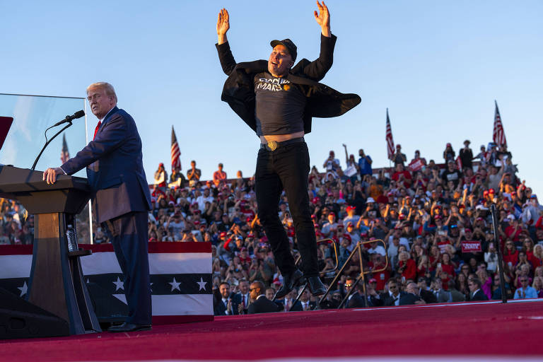 A imagem mostra um evento político ao ar livre, com uma grande multidão ao fundo. À esquerda, Donald Trump, de terno, está em um púlpito, aparentemente fazendo um discurso. À direita, Elon Musk, vestido com uma camiseta preta e calças escuras, está pulando e levantando as mãos, demonstrando entusiasmo. O cenário é decorado com bandeiras e há um tapete vermelho no chão.